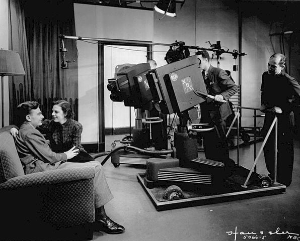 NBC Television In Black And White With Large Cameras Recording Two Actors Sitting On A Chair Looking At Each Other And Smiling