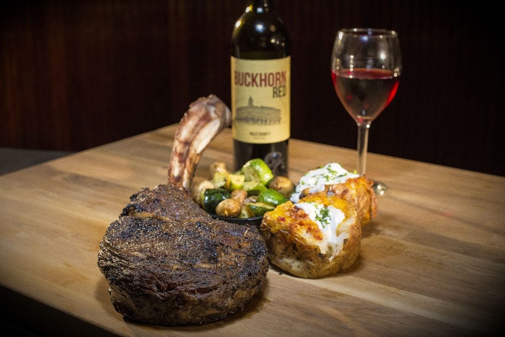a nicely lit image of a large piece of steak, a baked potato, and some Buckhorn Steakhouse wine in the background, all on top of a large wooden cutting board