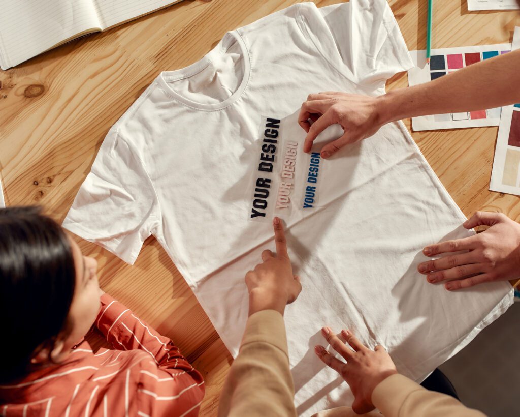 A truncated photo of three people working on a t-shirt design with a white t-shirt spread out on the table before them